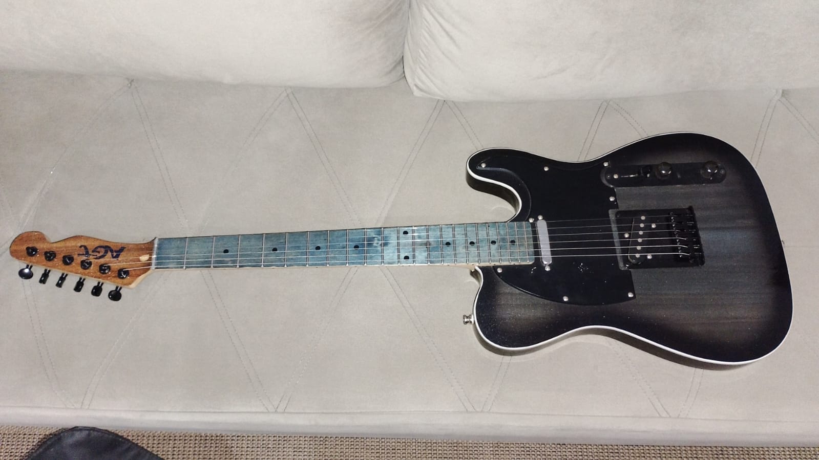 Guitarra AGT completa deitada sobre um sofá claro, mostrando corpo, braço e headstock